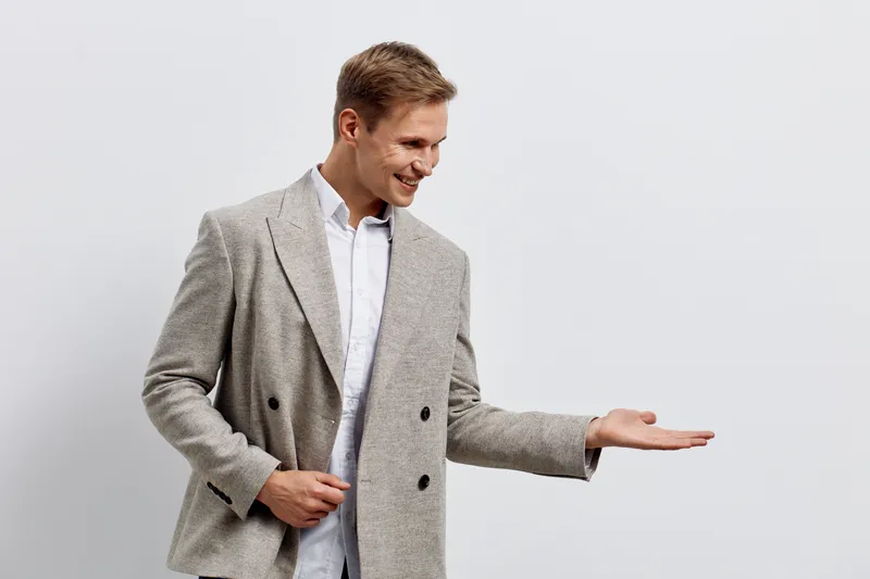 Confident young man in stylish gray blazer gesturing with hand on plain white background, smiling cheerful pose, casual outfit, modern fashion, studio portrait, positive mood, business casual style.