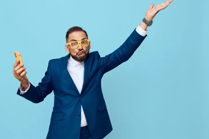 A professional businessman holds a smartphone and gestures upward against an isolated colored blue background, showing surprise and curiosity, signaling dynamic communication, confidence, and