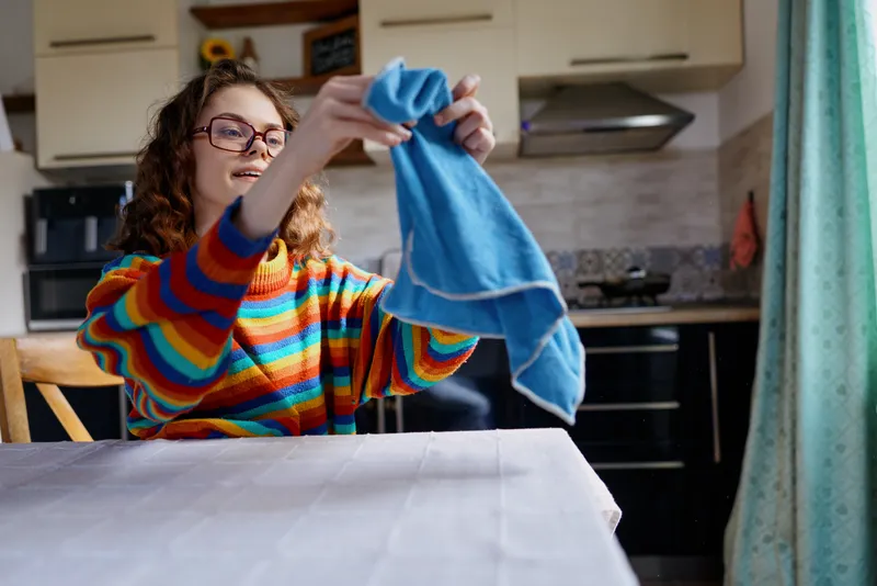 Cheerful woman indoors, drying clothes at a table, wearing a colorful striped sweater, casual home setting, bright natural light, relaxed mood, everyday domestic scene