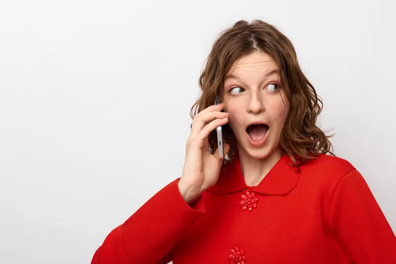 Surprised young woman with curly hair wearing red sweater talks on smartphone, looking aside with open mouth. Concept of unexpected news, communication, and emotional reaction.