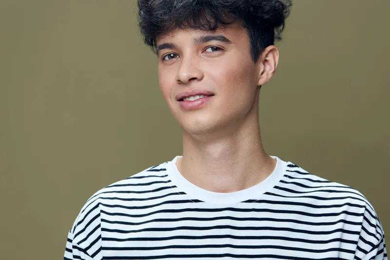 young man with curly hair wearing a striped shirt smiling softly against olive background with casual and natural expression, fresh and relaxed mood