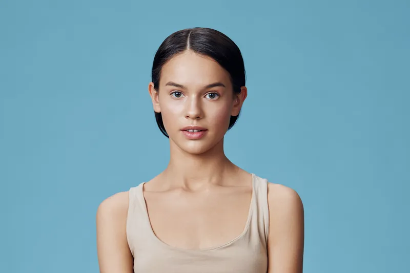 Young woman with short dark hair wearing a beige tank top against a light blue background, expressing calm and confidence, ideal for beauty and skincare concepts