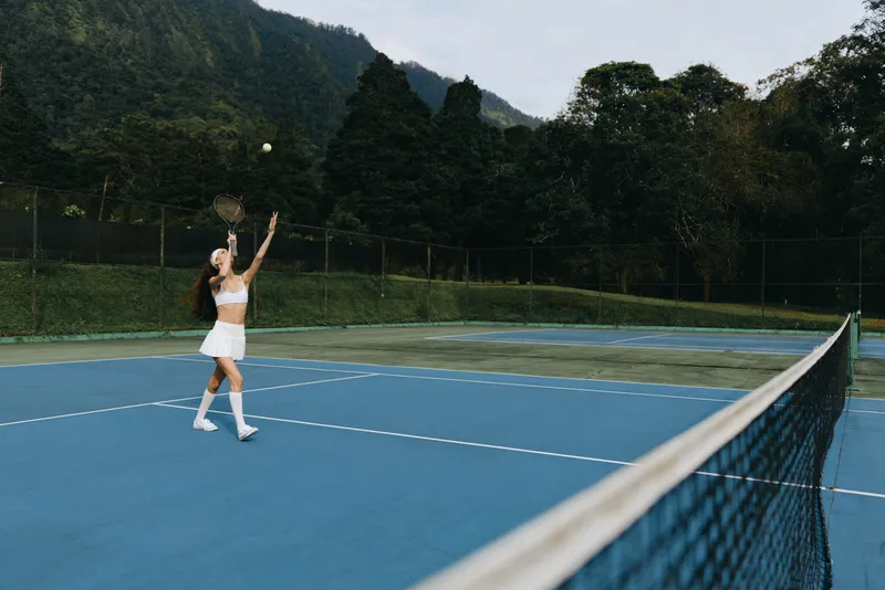 Female tennis player in a white sports outfit serving on a blue outdoor court surrounded by lush green mountains