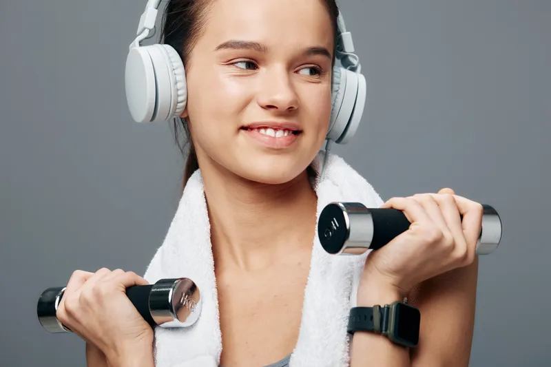 Fitness sport woman workout with headphones, lifting dumbbells and wrapping a towel over shoulders, captured during an energetic training moment in the gym.