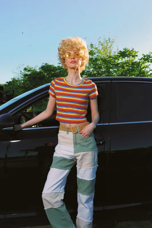Joyful woman outdoors in a striped crop top and patchwork denim, leaning by a dark car, sunlit breeze, retro film color, casual lifestyle and vibrant greenery behind in bloom today