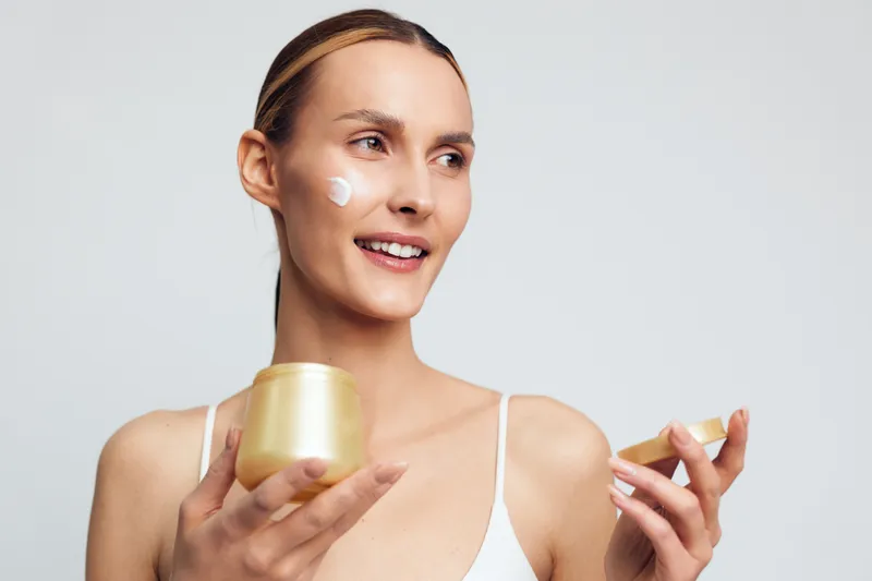 A smiling young woman applies face cream from a golden jar on her cheek, demonstrating skincare routine and natural beauty care with a clean light background and casual outfit.