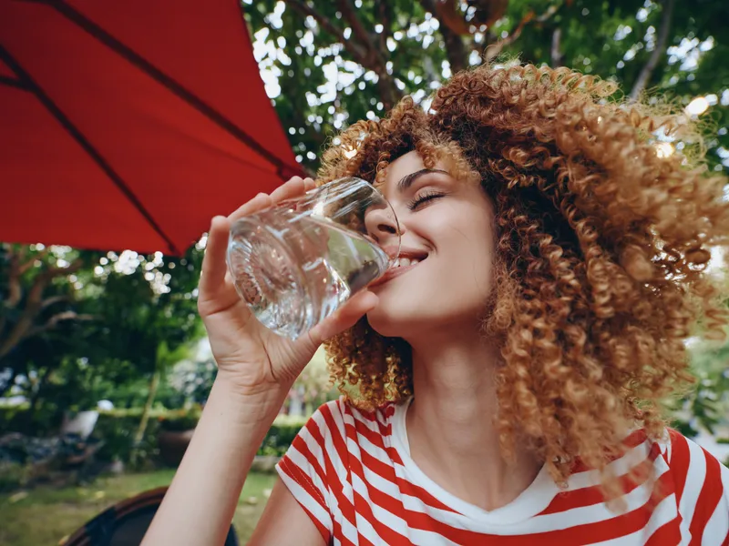 Woman with curly hair drinks water from a glass outdoors under a red umbrella, smiling and enjoying a refreshing moment on a sunny day in a park.