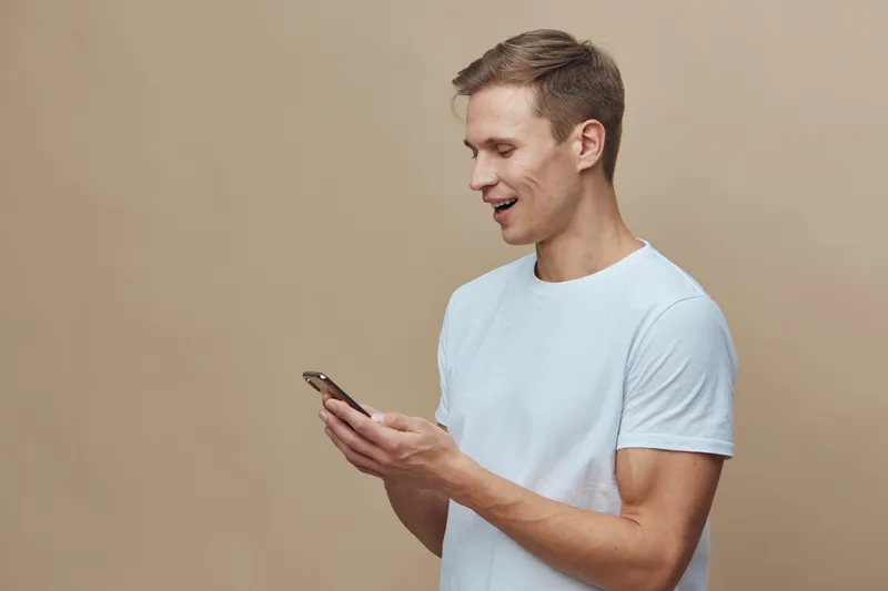 Smiling young man using smartphone in casual white t-shirt isolated on plain beige background studio portrait. People technology lifestyle concept