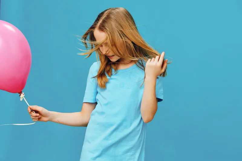 A young girl with blonde hair joyfully playing with a pink balloon against a vibrant blue background, capturing the essence of childhood innocence and happiness
