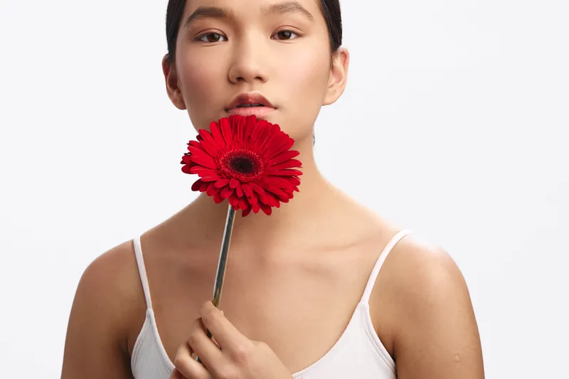 Asian woman gently holds a red flower near her lips in a bright studio setting, creating a calm, artistic portrait with soft shadows and elegant simplicity.