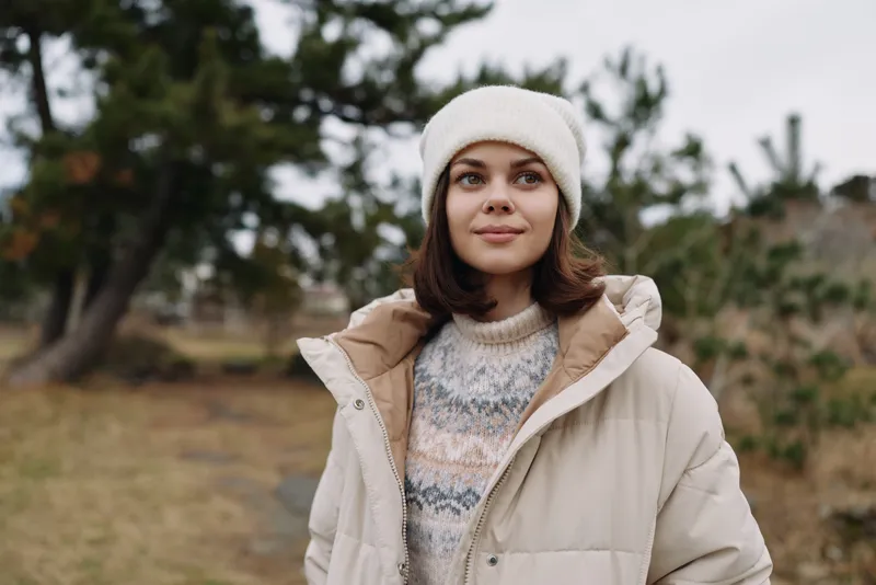 Woman outdoors in a beige winter coat and knit sweater, wearing a beanie, posing among trees in an open landscape with a serene, confident gaze