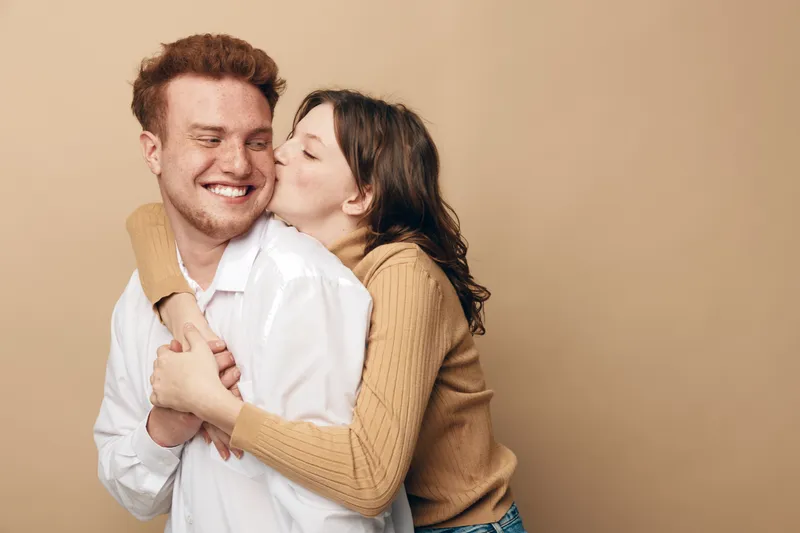 Happy young couple embraces and smiles in casual clothes on beige background. Woman kisses man on cheek showing affection and love. Concept of relationships, happiness, and intimacy.