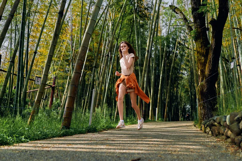 Woman running on a gravel path through tall bamboo forest, casual outfit with jacket tied at waist, outdoor exercise and joyful motion, summer nature scene with sunlight.