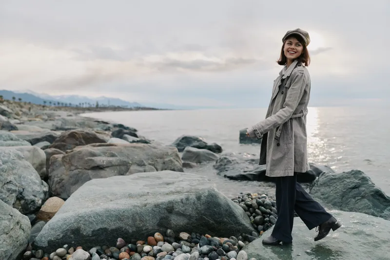 Young woman in coat and hat walks on rocky beach near calm sea under cloudy sky. Autumn coastal landscape with pebbles and distant mountains. Concept of outdoor lifestyle and nature.