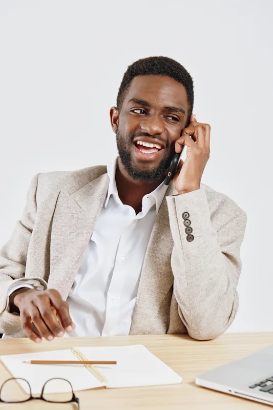 Smiling man talking on mobile phone in office setting, wearing beige blazer and white shirt, working desk with notepad, pen, and glasses, professional mood, indoor environment, natural light.