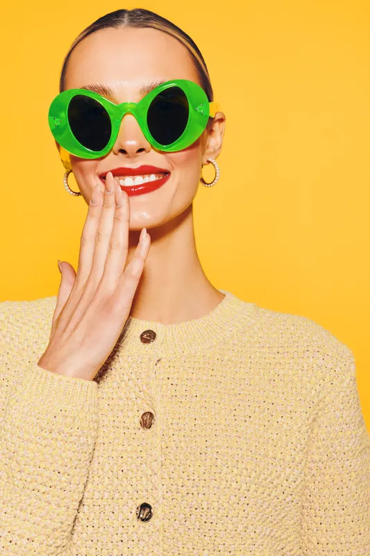 Young woman in bright green sunglasses and beige sweater smiles with hand near mouth on yellow background. Concept of fashion, style, summer, and joyful mood.