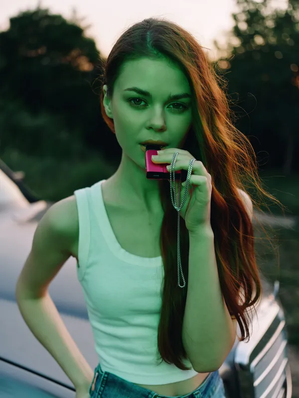 Young woman with long hair holding a charm, wearing a sleeveless top, with a serene expression against a natural sunset background, embodying youthful elegance and creativity.