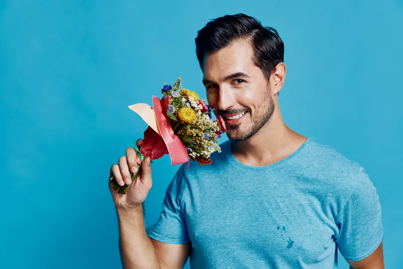 Handsome young man smiling while holding a colorful bouquet near his face, wearing a casual blue t-shirt, standing against a vibrant blue background. Floral lifestyle concept.