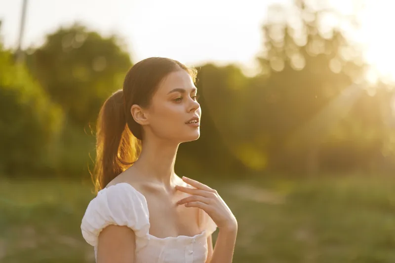 Young woman in a white dress enjoying a sunny evening, showcasing natural beauty and serenity in a lush green setting with warm sunlight