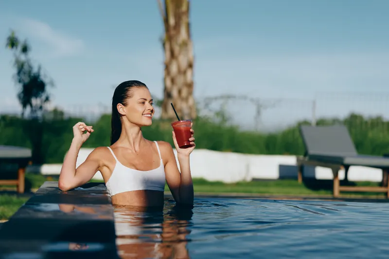 Young woman in white bikini relaxing in outdoor pool holding red drink, smiling, sunny day, calm water, natural background, leisure moment, healthy lifestyle concept