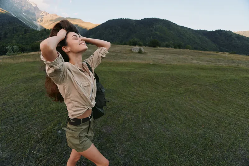 Woman enjoying outdoor life, standing in a meadow with arms raised, smiling, backpack visible. Nature mood, wellness, and freedom in a sunny landscape.