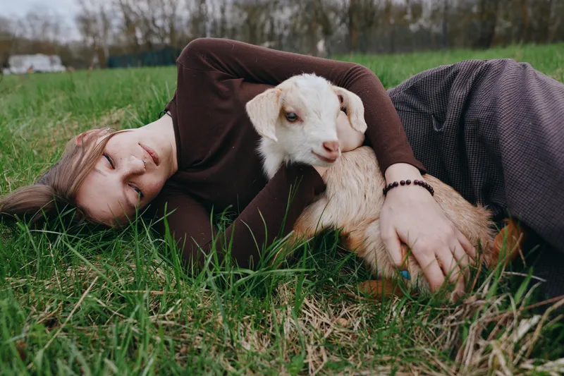 Goat kid cuddle scene featuring a relaxed woman lying on grass, embracing a gentle animal, showcasing lifestyle and outdoor calm, natural light, warm bond, candid countryside moment