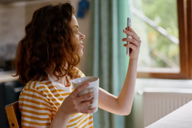 Woman with curly hair in a striped shirt sits at home, filming a selfie beside a window while holding a cup, capturing a casual morning routine and cozy self care mood.