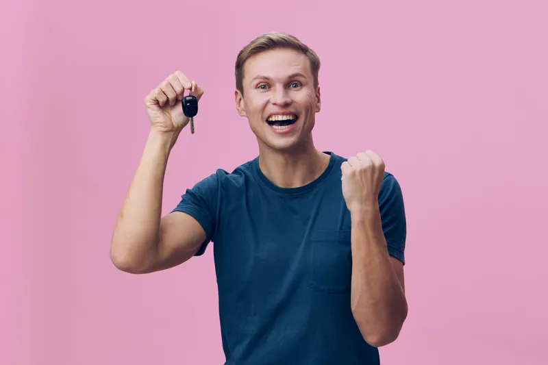 Smiling young man holding car key with excited expression on pink background, casual blue t-shirt, celebrating achievement, positive emotion, lifestyle concept.