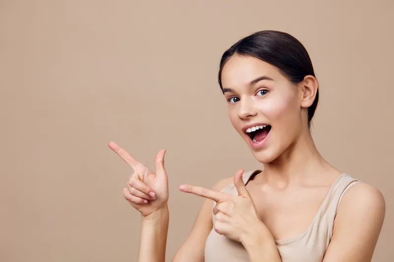 Happy young woman pointing to the sides with excitement on a neutral background, showcasing joy and enthusiasm in casual beige clothing