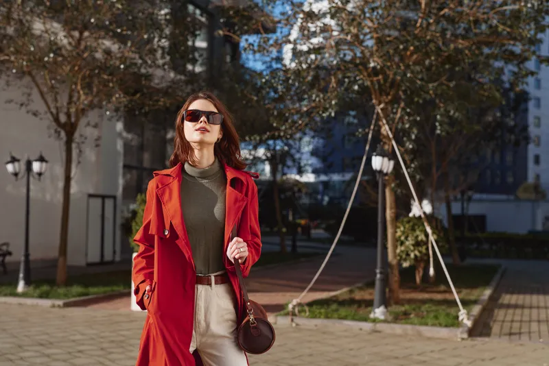 A stylish woman in a red coat walks along a sunny city street, wearing sunglasses and a brown bag, exuding confident urban style.
