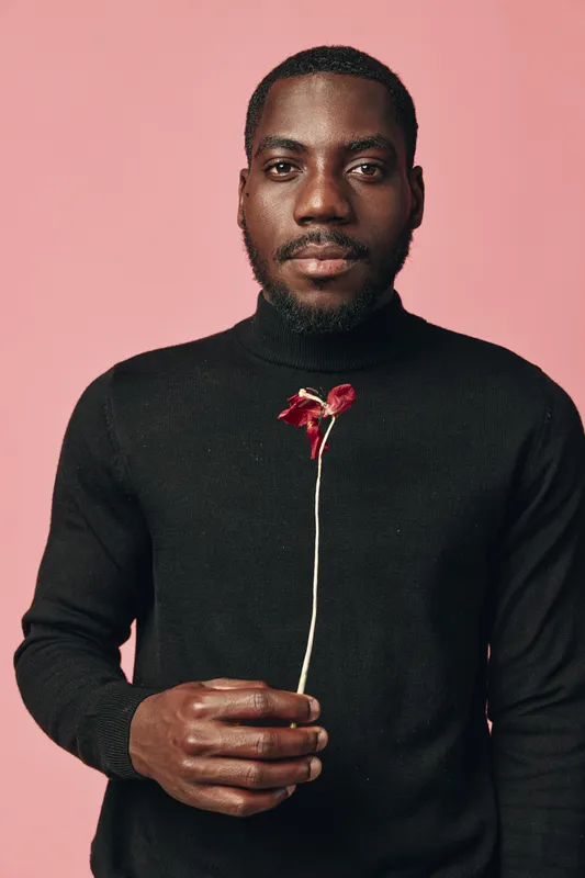 African man with dark skin wearing black sweater holding red flower on pink background creative conceptual visual style portrait with serious expression and minimalistic design.
