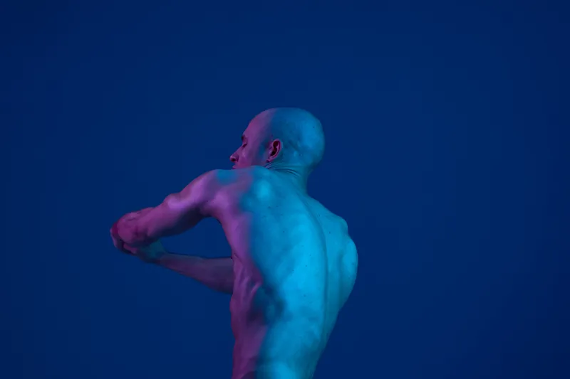 A man with a bald head is gracefully moving against a vibrant blue backdrop, showcasing the interplay of light and shadow. The colors create an artistic feel.