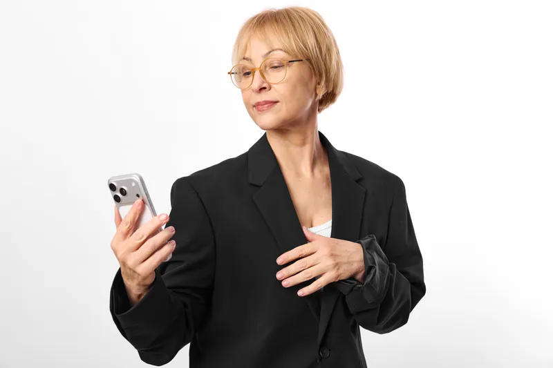 Woman in business suit holds smartphone for communication, calm mature office portrait, glasses visible, professional gesture, clean studio light, white background