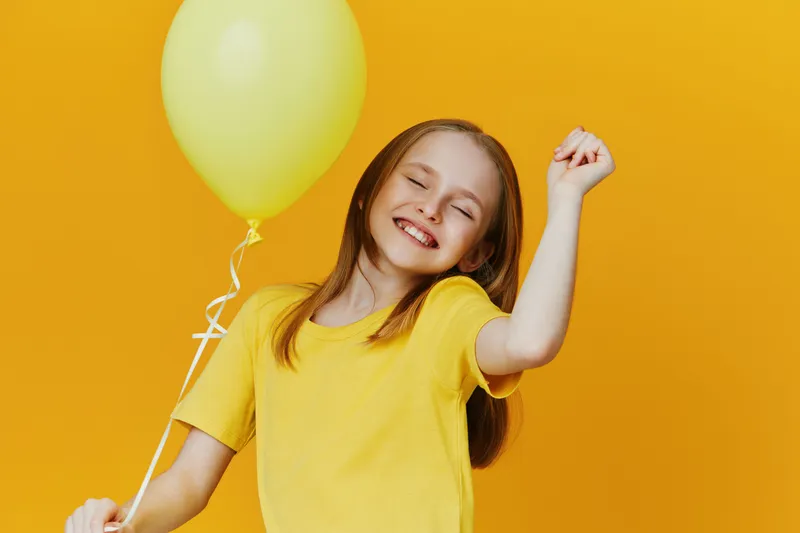 Happy girl in a yellow t shirt holding a yellow balloon, celebrating joyfully against a bright yellow background, perfect for concepts of happiness and childhood