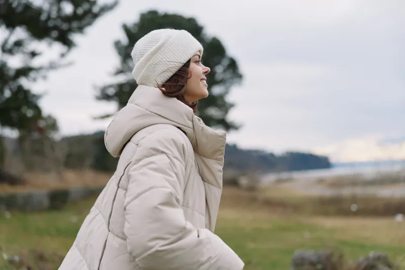 Winter scene with a woman in a warm beige coat and knit beanie, smiling as she gazes toward a sprawling landscape of fields and trees under a soft cloudy sky