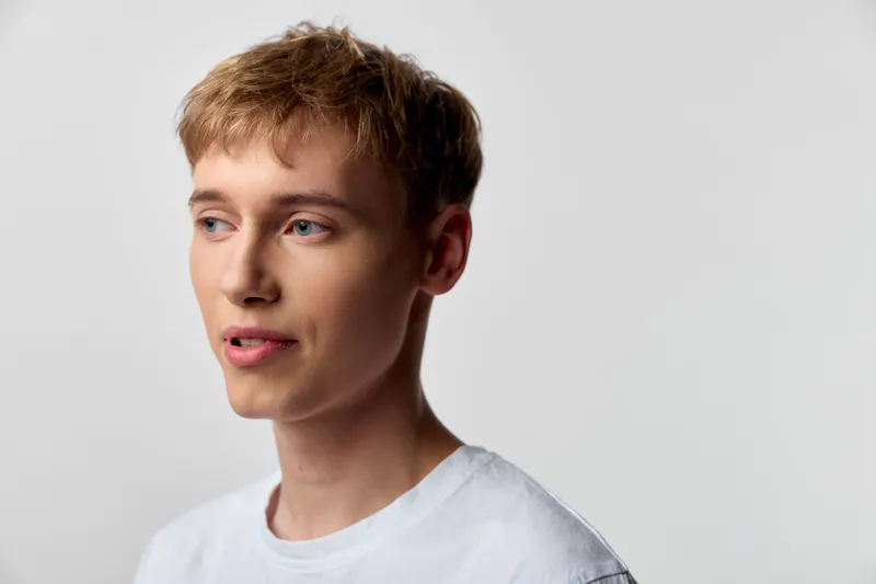 Young man in casual attire wearing a white shirt stands against a neutral studio backdrop, looking aside with a thoughtful expression, ideal for lifestyle, fashion, and marketing visuals.