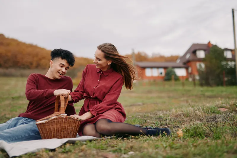 A genuine candid picnic scene of a young couple sharing laughter as they reach into a wicker basket on a blanket in an autumn meadow, capturing natural connection, trust and authenticity.