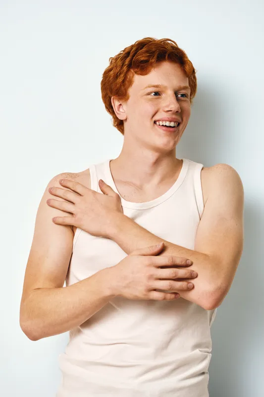 Young man with red hair and fair skin wearing white tank top smiling and looking away while crossing arms, isolated on white background with casual posture and positive expression.