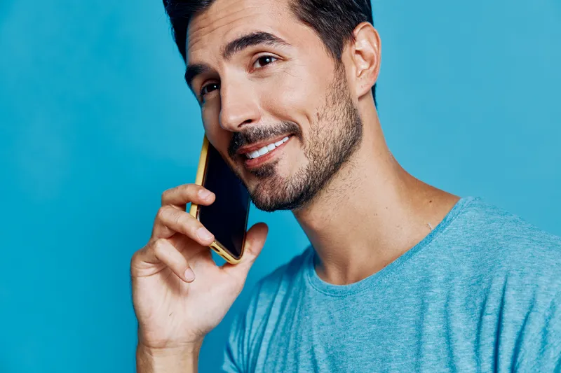 Smiling young man with stylish beard talking on smartphone, wearing casual blue t-shirt, isolated on solid turquoise background. Communication and lifestyle concept.