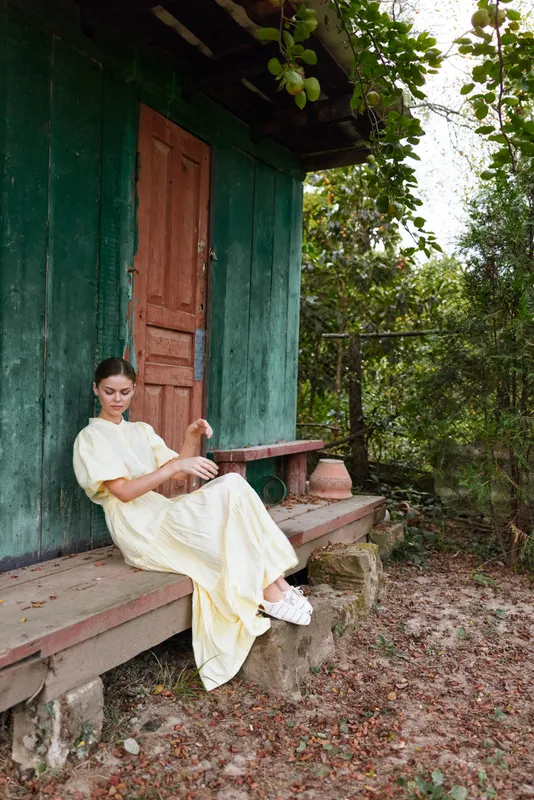 A woman in a pale yellow dress sits on a wooden porch beside a green rustic cabin, amid trees and fallen leaves, creating a serene countryside mood and gentle outdoor charm.