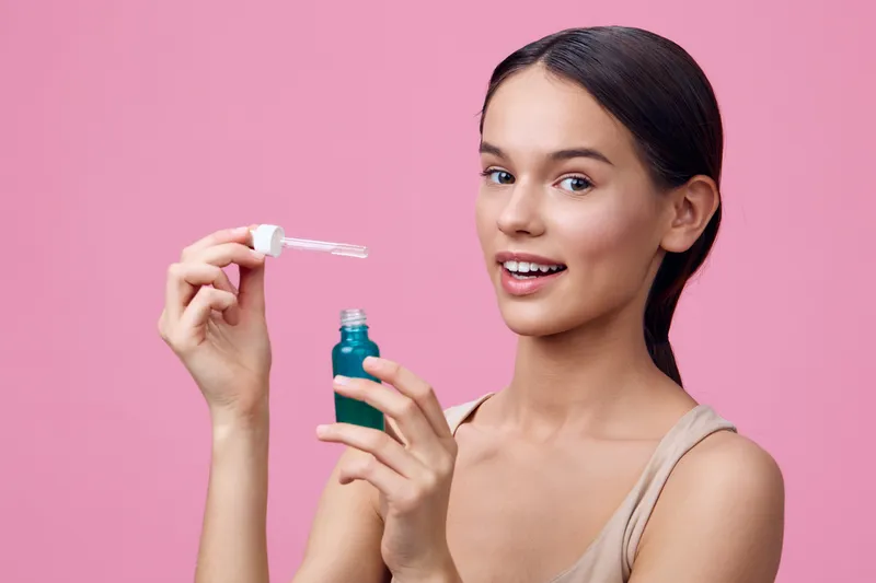 Young woman applying serum, showcasing healthy skin and natural beauty, on a pink background, conveying joy and confidence in skincare routine