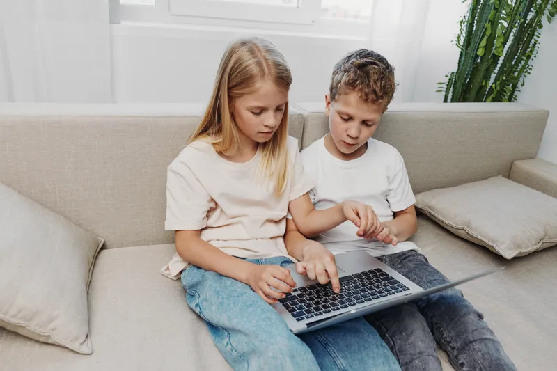 Two children sitting on a sofa using a laptop together, focused on the screen. Concept of learning, technology, collaboration, and childhood education at home.