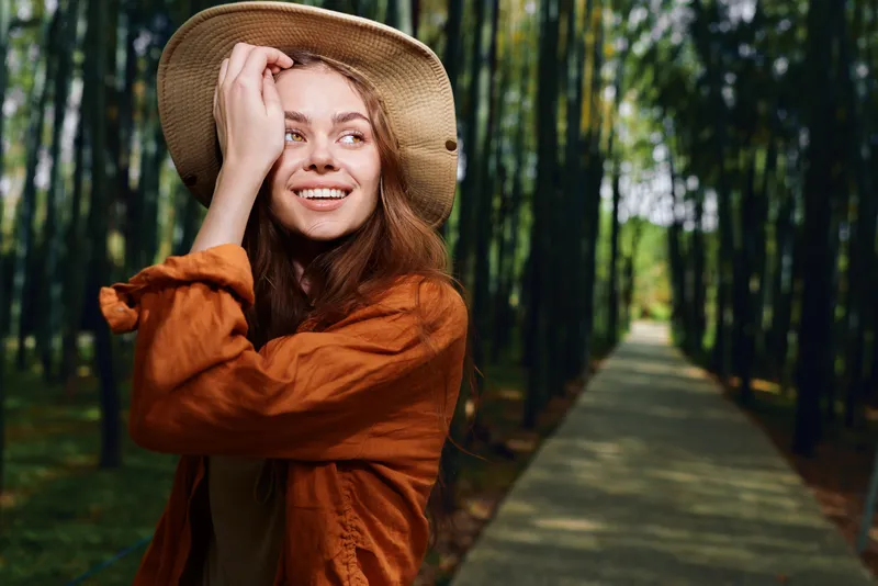 Woman smile hat park walkway portrait outdoors nature, young woman in a straw hat smiling on a tree-lined path, casual fashion and sunlight on face, joyful relaxed lifestyle and travel mood.