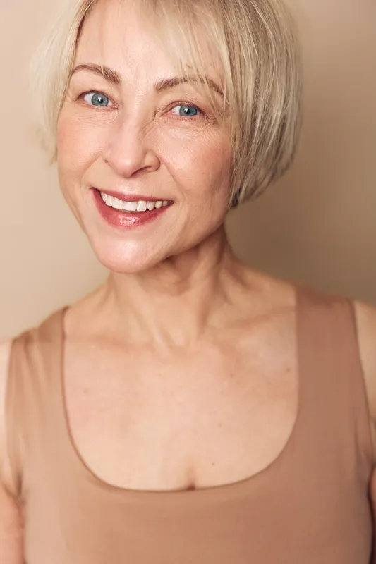 Smiling mature woman with short blonde hair and blue eyes wears beige top, showing natural skin and joyful expression. Portrait in soft light with neutral background for beauty and aging concept.