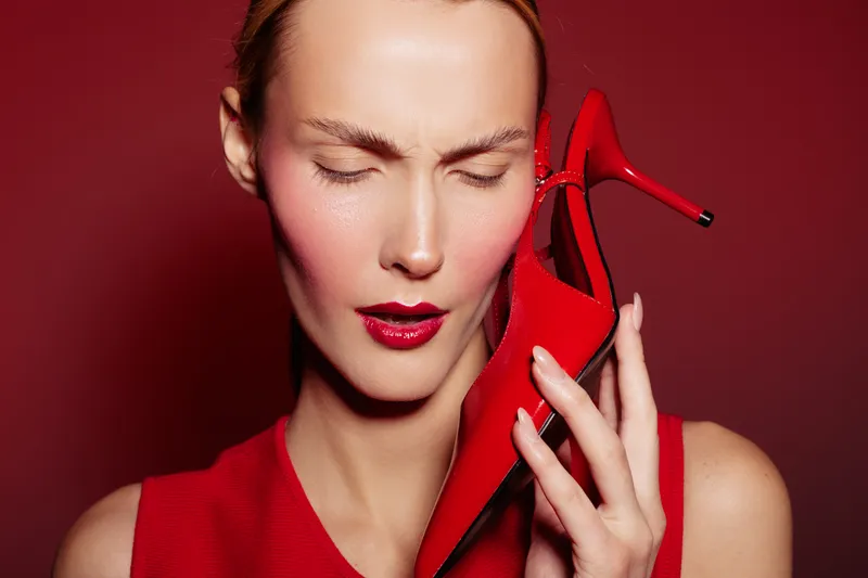 Woman in red dress holds red high heel shoe near face with closed eyes on red background. Concept of fashion, style, elegance, and bold color contrast in studio lighting.