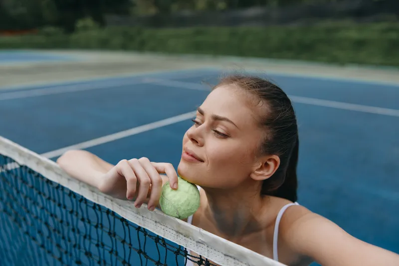Young female tennis player enjoying a moment on the court with a tennis ball, showcasing emotions of relaxation and joy, surrounded by a green outdoor background