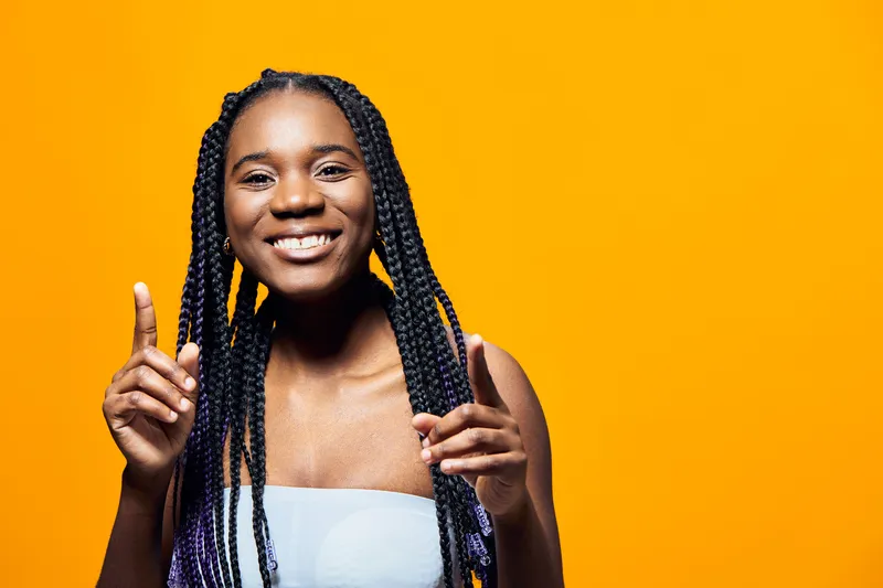 Smiling young Black woman with braided hair, wearing a casual top, gesturing happily against a vibrant yellow background, conveying joy and positivity