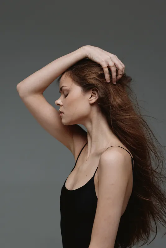 thoughtful woman with curly hair in black top posing against gray background, calm and elegant atmosphere