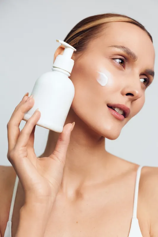 Young woman with blonde hair applies white cream on her cheek while holding a white lotion bottle near her face against a light background.
