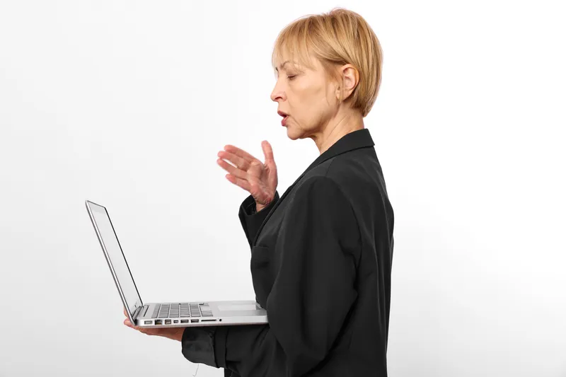 Woman with laptop in office, profile speaking gesture in studio, white background, blond adult standing, focused, hand raised while typing quietly